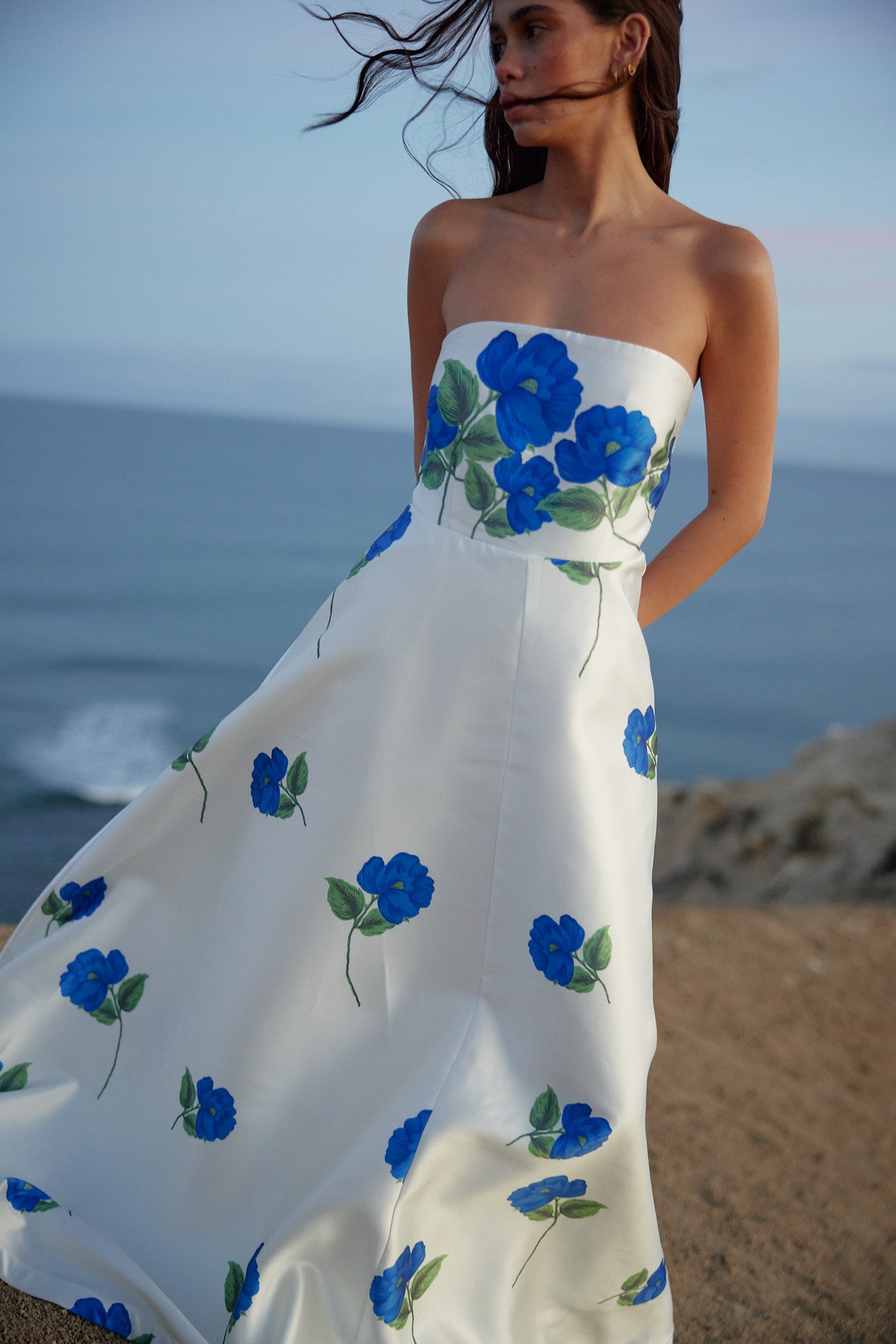 Woman wearing a strapless white dress with blue floral patterns by the ocean.