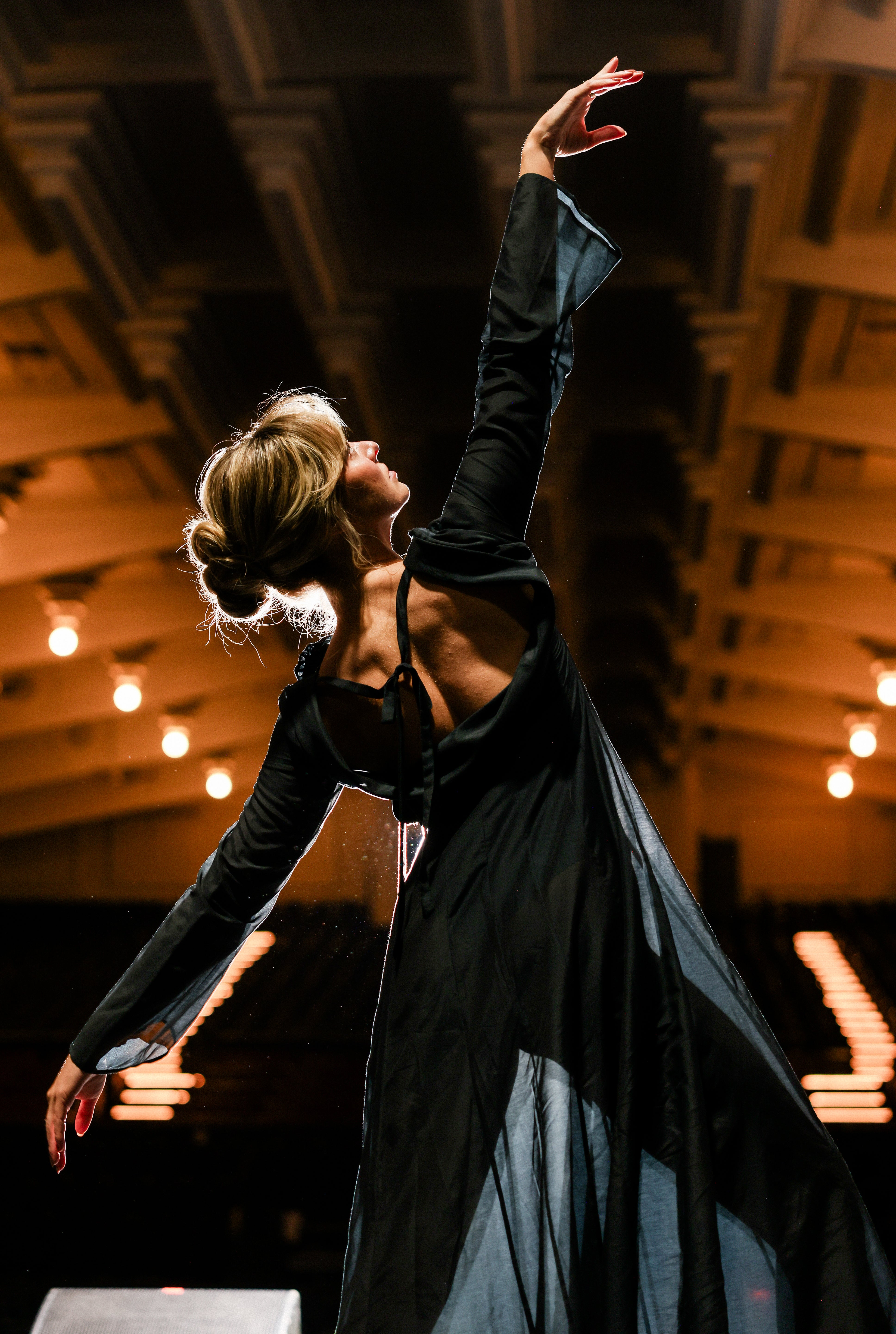 Dancer in the Odile Gown, a sheer black long sleeve evening gown, arms raised on stage